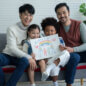 happy asian young lgbtq gay couple with little cute adopted caucasian and african kid smiling and showing rainbow family drawing in living room at home. lgbt diverse family concept