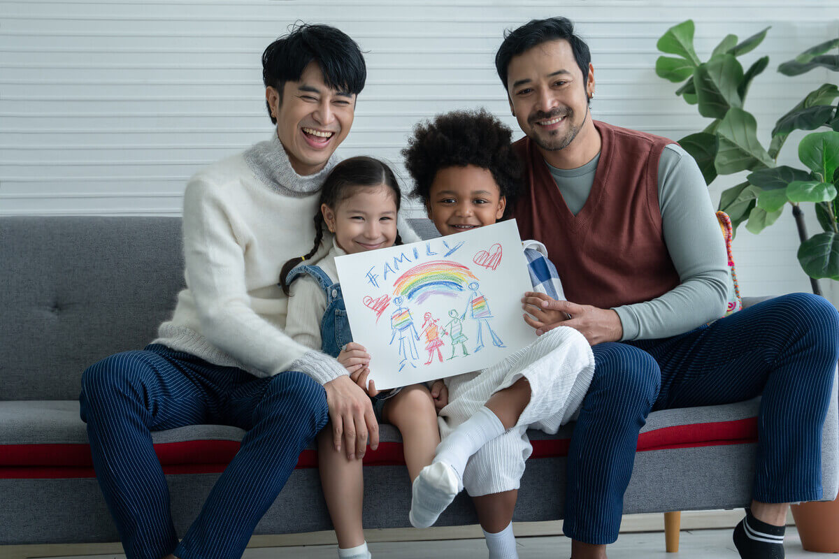 happy asian young lgbtq gay couple with little cute adopted caucasian and african kid smiling and showing rainbow family drawing in living room at home. lgbt diverse family concept